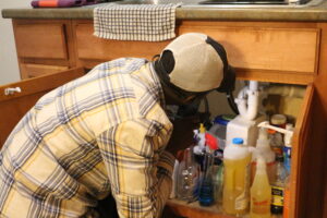 A city employee inspecting the plumbing under a kitchen sink inside a resident's home.