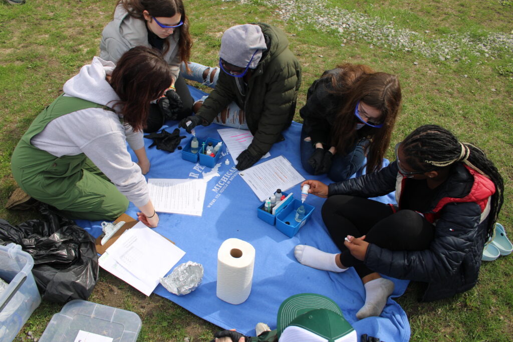 Students from Flint Southwestern Academy sit on a blanket with testing equipment to measure the dissolved oxygen levels of Flint River water in the Mott Park Recreational Area. 