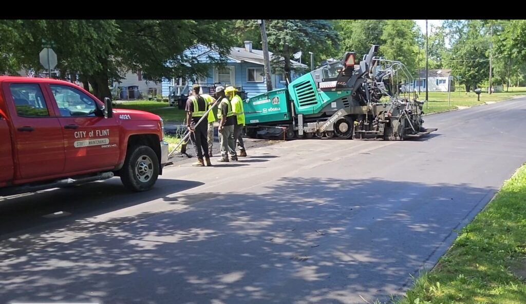 Street maintenance workers near a city truck and a paver on a freshly paved neighborhood street