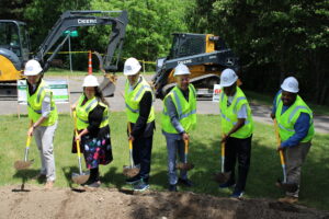 Mayor Sheldon Neeley, former Congressman Dan Kildee, and other stakeholders pose in hard hats, reflective vests, and shovels in front of a mound of soil at the site of the St. John Street Neighborhood Memorial Park with construction equipment behind them.