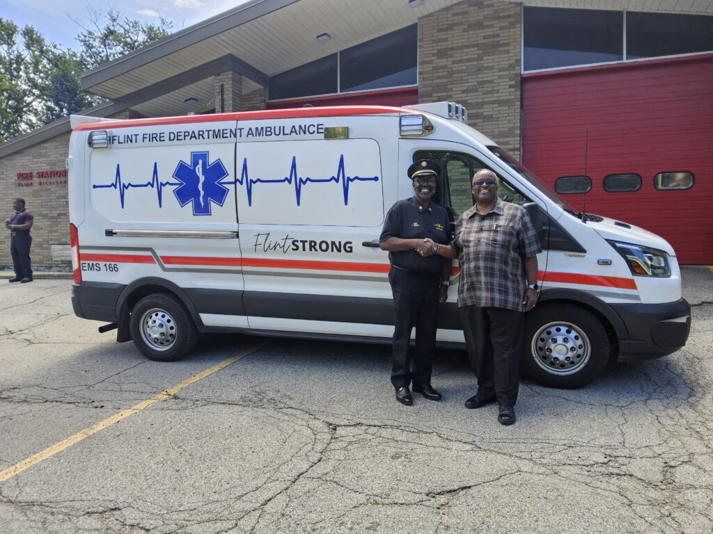 (From left) Flint Fire Chief Theron Wiggins and City Administrator Clyde Edwards shaking hands in front of a City Fire Department ambulance parked in front of Flint Fire Station #8.