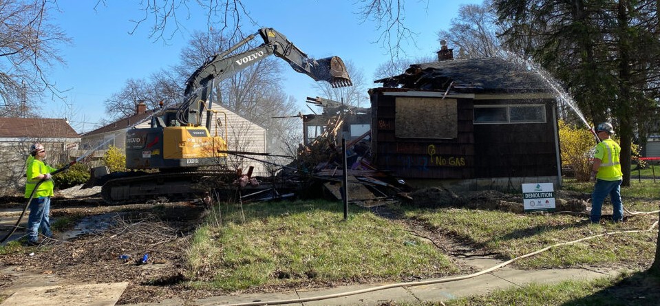 The demolition of a house underway with half of the structure already razed by an excavator while two workers hose the site down with water to mitigate dust spread.